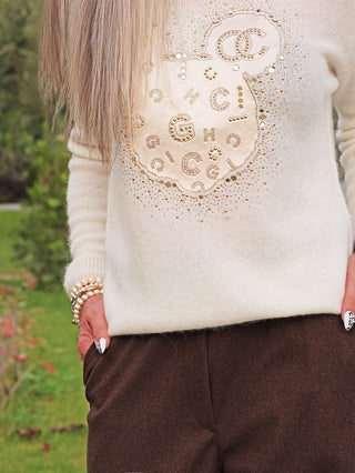 OFF#DLY Pulli Kira white zeigt einen cremefarbenen Strickpullover mit goldenen Nieten und Strasssteinen in einem kreisförmigen Muster, getragen mit dunklen Hosen und einer Perlenarmband.