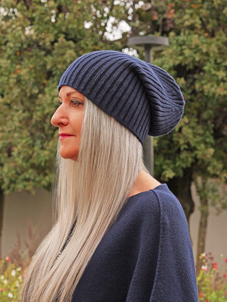 Frau trägt eine OFF#DLY Mütze Anna marine, ein dunkelblaues, geripptes Strick-Beanie, passend zu ihrem Outfit. Sie steht in einem herbstlichen Garten mit langen, silbergrauen Haaren.