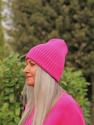 Eine Frau trägt die OFF#DLY Mütze Anna pink, eine leuchtend pinke Strickmütze, passend zu ihrem pinken Oberteil, vor einem grünen, blattreichen Hintergrund.