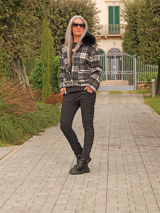 Person trägt OFF#DLY Jacke Checker black/grey mit abnehmbarem Kunstfellkragen, karierter Oberfläche und seitlichen Taschen, spaziert auf einem Gartenweg vor einem verzierten Tor mit gepflegter Landschaft.