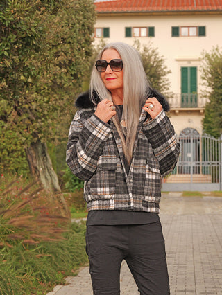 Eine Frau trägt die OFF#DLY Jacke Checker black/grey mit Karomuster, Kunstfellkragen und kurzem Schnitt, kombiniert mit schwarzen Hosen, vor einer Villa.