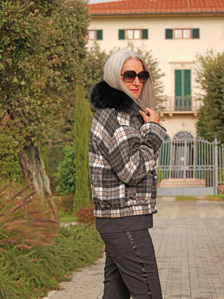 Frau trägt OFF#DLY Jacke Checker black/grey mit schwarzem Kunstfellkragen und Karomuster, stehend vor einer Villa. Die Jacke hat einen lässigen, kastigen Schnitt, ideal für kühle Tage.