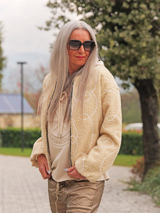 Erwachsene Frau trägt die OFF#DLY Jacke Alina beige, eine kastige Teddyjacke mit Reißverschluss, in neutralem Ton. Perfekt für kühle Tage, kombiniert mit hellen, glänzenden Jogginghosen draußen im Park.