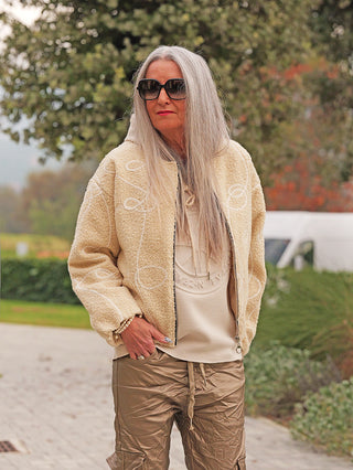 Ältere Frau trägt die OFF#DLY Jacke Alina beige, eine bequeme Fleecejacke im Teddy-Look mit Reißverschluss, kombiniert mit einer Kapuze und seitlichen Taschen, ideal für kühle Tage.