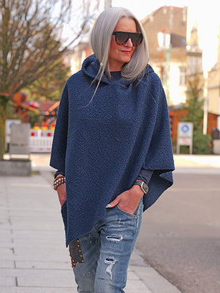 Person trägt den OFF#DLY poncho Teddy blue, ein locker sitzendes, strukturiertes, navy-blaues Kapuzen-Poncho aus weichem Stoff, kombiniert mit einer langärmligen Oberteil und gepatchten Jeans.