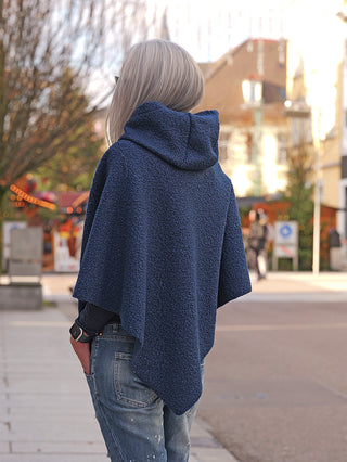 Eine Person trägt den OFF#DLY Poncho Teddy blue, einen strukturierten, navy-blauen Kapuzenponcho, der locker über einen dunklen Pullover und hellblaue Jeans fällt, während sie auf einer gepflasterten Straße steht.