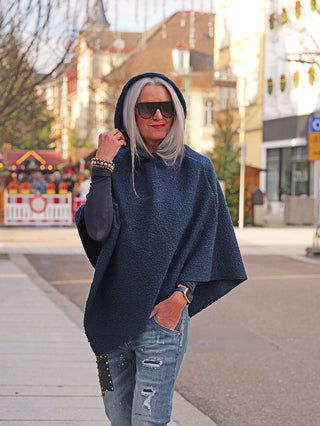 Eine Frau trägt den OFF#DLY poncho Teddy blue, einen dunklen, texturierten Kapuzenponcho, über einem Langarmshirt, kombiniert mit zerrissenen Jeans, und steht auf einer belebten Straßenszene.