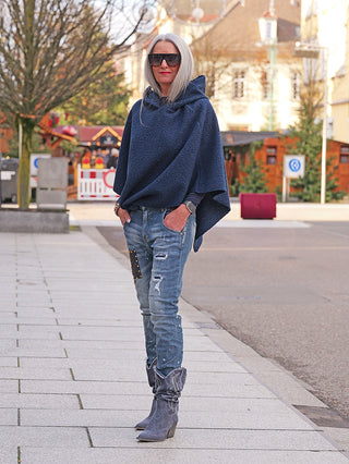 Frau in Destroy Denim Dora Jeans mit Used-Waschung, Farbeffekten und Patch über Knie, kombiniert mit grauen Cowboy-Stiefeln und navy-blauem Poncho, präsentiert lässigen, modernen Streetstyle.