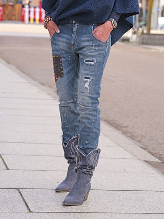 OFF#DLY Destroy Denim Dora midblue Jeans mit schmalem Schnitt und Used-Look, inklusive kleinen Rissen, Farbspritzern und einem nietenbesetzten Patch, getragen mit grauen Wildleder-Cowboy-Stiefeln.