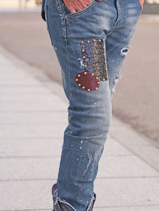 OFF#DLY Destroy Denim Dora midblue – Nahaufnahme: Beine in schmal geschnittenen, distressed Jeans mit zwei dekorativen Patches und Nieten; Used-Look mit Farbspritzern, Hand in der Tasche.