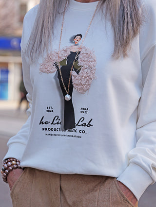 Nahaufnahme des OFF#DLY Sweat Laura white mit einem modischen Print einer stilisierten Frau im schwarzen Kleid und pinkem Wrap, kombiniert mit einer goldenen Perlenkette und braunen Wildlederhosen.