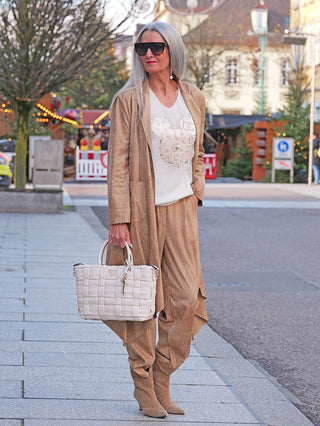 Frau trägt den OFF#DLY Mantel Velourstoff Gardone camel, locker sitzend, mit fließendem asymmetrischen Schnitt, über einem Sweater, in Kombination mit passenden weiten Hosen und strukturiertem Satchel.