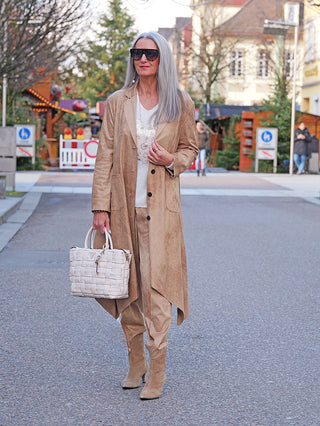 Eine silberhaarige Frau trägt den OFF#DLY Mantel Velourstoff Gardone camel in fließendem, asymmetrischem Schnitt, kombiniert mit passenden tanfarbenen Stiefeln und einer gewebten Tasche.