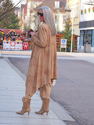 Frau trägt OFF#DLY Mantel Velourstoff Gardone camel, ein langer, fließender Mantel in Veloursleder-Optik. Kombiniert mit passenden Hosen und Stiefeln, vermittelt er Eleganz im winterlichen Marktszenario.