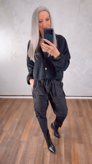 Person mit langen, silbernen Haaren im Spiegelbild trägt Thom Krom Hose MP25 black. Die Cargo-Hose hat einen elastischen Bund mit Drawstring und auffällige Taschen, kombiniert mit schwarzen Stiefeln.