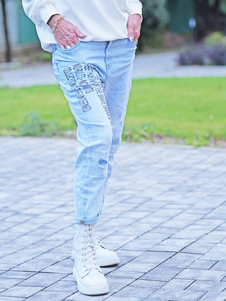 Person trägt OFF#DLY Denim Destroy Thea lightblue Jeans mit vertikalem Schriftprint und leichten Destroy-Details. Die Jeans sind hochgekrempelt, kombiniert mit weißen High-Top-Sneakers auf einem gepflasterten Platz.