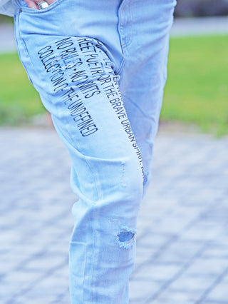 Nahaufnahme von OFF#DLY Denim Destroy Thea lightblue Jeans mit schwarzem Schriftzug am Oberschenkel, leicht zerstörten Details am Knie und einer Hand mit klobigem Ring in der Tasche.