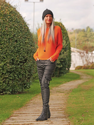 Person in einem OFF#DLY Pulli Cara orange, einem modernen, gerippten, leicht verkürzten Pullover, kombiniert mit schwarzen Cargo-Hosen, schwarzen Stiefeletten und einer dunklen Mütze, stehend auf einem Gartenweg.