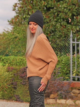 Person im Garten trägt den OFF#DLY Pulli Cara camel, ein locker geschnittenes, geripptes, tanfarbenes Oberteil, kombiniert mit dunklen Hosen und einer schwarzen Mütze, umgeben von Pflanzen und Zaun.