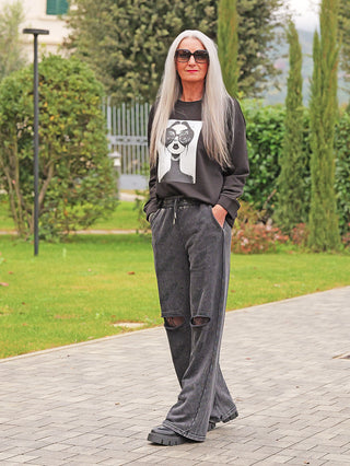 Person trägt die OFF#DLY Joggingpant Glitzer Destroy black mit weitem Schnitt, glitzernden Akzenten und Destroy-Details an den Knien, kombiniert mit schwarzen Plattform-Sneakern auf einem gepflasterten Weg.