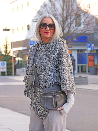 Eine Frau trägt ein OFF#DLY Poncho Teddy salt/pepper, ein grob gestricktes, graues Cape mit Kapuzenstil. Dazu kombiniert sie fingerlose Handschuhe und eine strukturierte Umhängetasche.