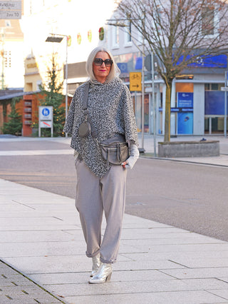 Eine Person trägt ein OFF#DLY Poncho Teddy in Salz/Pfeffer-Optik. Der Poncho ist grob gestrickt und ergänzt das graue Ensemble aus lockerer Hose und silbernen Stiefeletten auf einem städtischen Bürgersteig.