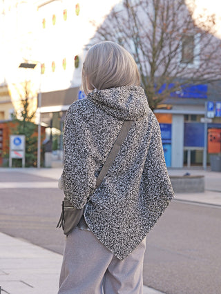 Person von hinten, trägt den OFF#DLY Poncho Teddy salt/pepper über grauer Hose, mit einer kleinen grauen Umhängetasche. Sie geht eine gepflasterte Stadtstraße entlang, umgeben von Geschäften und kahlen Bäumen.