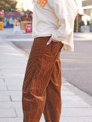 OFF#DLY Cord Pant Tabea camel getragen von einer Person auf dem Gehweg, kombiniert mit einem cremefarbenen Sweatshirt. Weit geschnittene, braune Cordhose, hand in der Gesäßtasche, zeigt schwarz-weiß gemusterte Nägel.