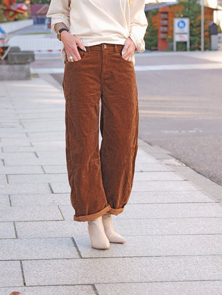 OFF#DLY Cord Pant Tabea camel: Locker sitzende, hochgekrempelte braune Cordhose, getragen mit Händen in den Taschen, zeigt Vintage-Touch und lässigen Stil, kombiniert zu cremefarbenen Stiefeletten.