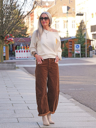 Frau in OFF#DLY Cord Pant Tabea camel, weite, lässige Passform. Kombiniert mit einem cremefarbenen Oberteil und beigen Stiefeletten. Ideal für einen entspannten, stylischen Stadtlook.