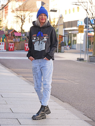 Person mit dem OFF#DLY Sweat DOG black Hoodie, das Hunde mit Zeitungen zeigt. Sie trägt eine blaue Mütze, Jeans und schwarze Stiefel, stehend auf einer städtischen Straße.