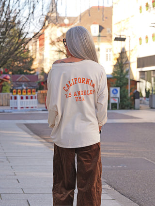 Eine Person trägt den OFF#DLY Sweater LA creme/orange mit lockerem Schnitt und „California Los Angeles USA“-Print auf dem Rücken, kombiniert mit weiten braunen Hosen auf einem städtischen Gehweg.