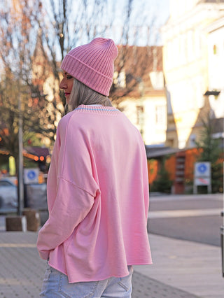 Person von hinten in einem OFF#DLY Sweater Lecco rose, locker sitzend mit rundem Ausschnitt. Kombiniert mit einer rosa Strickmütze und Jeans, in einer Fußgängerzone stehend.