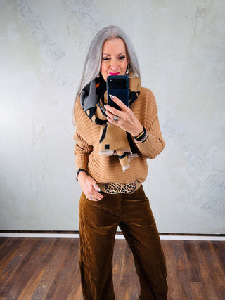 Person mit langem grauem Haar trägt den OFF#DLY Schal C.C. black/beige mit grafischem Muster. Der Schal ergänzt einen camel-farbenen Pullover und braune Cordhose in einem schlichten Raum.