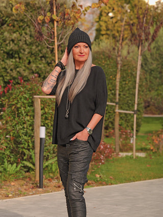 Person trägt den OFF#DLY Pulli Angelina in Schwarz mit halblangen, gerippten Ärmeln und lockerem Schnitt, kombiniert mit schwarzen Hosen und Beanie, in einem parkähnlichen Umfeld.