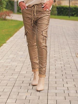 OFF#DLY Joggpant Bella gold: Metallic-beige Cargo-Hose mit Kordelzug und Klapptaschen, getragen mit beigefarbenen, knöchelhohen Stiefeln auf einem gepflasterten Weg, betont durch edlen Glanz und Streetstyle.