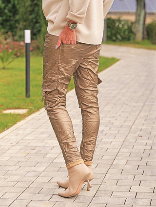 Person trägt die OFF#DLY Joggpant Bella gold mit seitlichen Cargo-Taschen und Zippern, kombiniert mit spitzen Stiefeletten und einer cremefarbenen Pullovermanschette, während sie auf einem Weg geht.