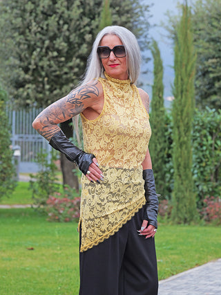 Frau im Park trägt das OFF#DLY Spitzen Top gelb, ein ärmelloses, gelbes Spitzentop mit asymmetrischem Saum und hohem Kragen, kombiniert mit schwarzen Hosen und fingerlosen Lederhandschuhen.