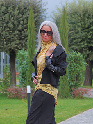 Frau trägt ein OFF#DLY Spitzen Top gelb mit hohem Kragen und asymmetrischem Saum, kombiniert mit schwarzen Accessoires, darunter eine Jacke, weite Hose und Lederhandschuhe, in einem gepflegten Garten.