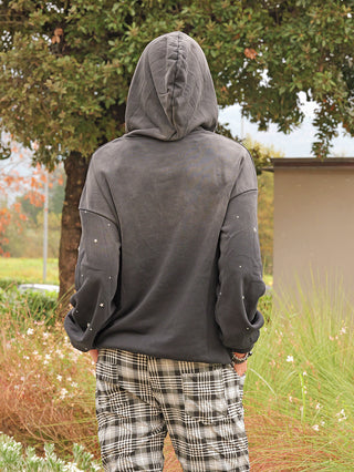 Person von hinten in einem Garten, trägt den OFF#DLY Hoody Pia black washed mit kleinen Stern-Nieten auf den Ärmeln und eine Kapuze, kombiniert mit schwarz-weiß karierten Hosen.