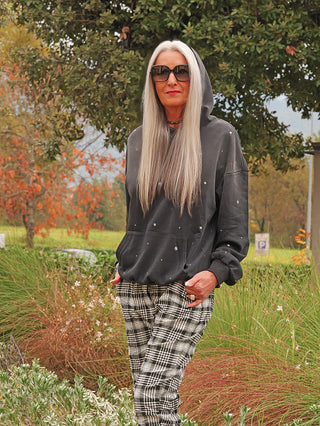 Ältere Frau trägt OFF#DLY Hoody Pia black washed mit Stern-Nieten und Punktdetails, präsentiert lässigen, verwaschenen Look in Parkumgebung. Lockerer Schnitt mit Kapuze und Kängurutasche.