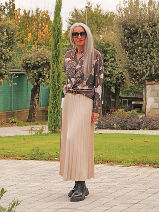 Frau trägt den OFF#DLY Plissee Rock beige, einen fließenden, knitterarmen Rock mit feinen Falten, kombiniert mit einem schwarzen Gürtel, passend zu ihrer stilvollen Bluse und robusten Stiefeln.