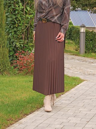 Person trägt den OFF#DLY Plissee Rock coffee, einen fließenden, schokoladenbraunen Faltenrock, gestylt mit einem schwarzen Gürtel und beigen Stiefeletten, auf einem gepflasterten Weg.