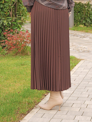 Person trägt den OFF#DLY Plissee Rock coffee auf einem Gehweg. Der Rock zeigt eine fließende Silhouette und feine Falten, kombiniert mit creme-beigen, spitzen Ankle-Boots.