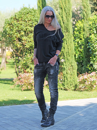 Frau trägt OFF#DLY Denim Julia Black Wash, schwarze Jeans mit Destroy-Details und lässigem Schnitt, kombiniert mit schwarzem, schulterfreiem Top und Ankle Boots, in einem Garten spazierend.