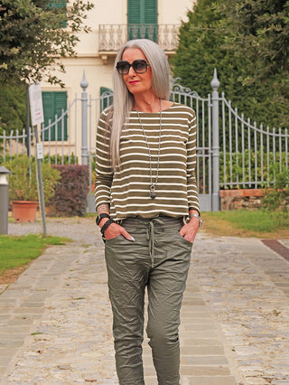 Mature Frau mit silbrig-grauem Haar in Streifenshirt OFF#DLY Ringel Donna khaki, lässig geschnitten, geht auf einem Gartenweg. Sie trägt Sonnenbrille, olivefarbene Hose und eine lange Halskette.