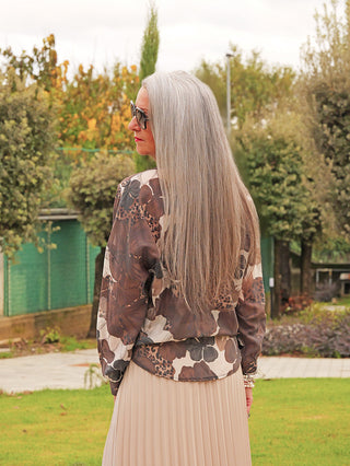 Frau im Garten trägt die OFF#DLY Bluse Tanja braun/mix mit abstraktem Muster, lockerem Schnitt und klassischem Kragen. Kombiniert mit beigem Rock, sorgt sie für einen eleganten Look.