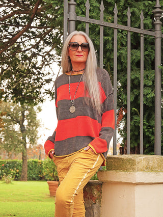 Person im Freien lehnt an Steinmauer, trägt OFF#DLY Pulli Bonny red/black mit breiten Streifen und lockerem Schnitt, kombiniert mit Sonnenbrille und Halskette für entspannten, modischen Look.