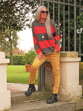 Ältere Frau trägt den OFF#DLY Pulli Bonny red/black mit breiten Streifen, kombiniert mit schmalen gelben Hosen und schwarzen Stiefeln, stehend vor einem schmiedeeisernen Tor in einer Gartenumgebung.