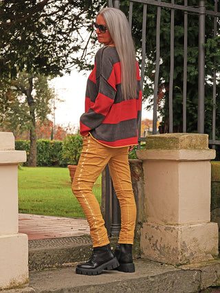 Person trägt den OFF#DLY Pulli Bonny red/black mit breiten horizontalen Streifen, kombiniert mit schmalen, gemusterten Hosen und robusten Schnürstiefeln, vor einem herbstlichen Parkeingang.